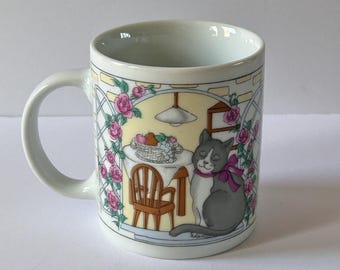 May include: White ceramic mug with a colorful illustration of a cat sitting at a table with a bowl of fruit. The cat is gray and white with a pink bow. The background is a yellow and white kitchen scene with pink roses.