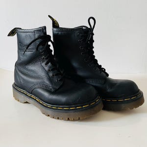 Może przedstawiać: Para czarnych skórzanych butów ze sznurowaniem. Buty mają grubą, bieżnikowaną podeszwę z żółtymi przeszyciami i uchwytem u góry. Buty mają około 15 cm wysokości.