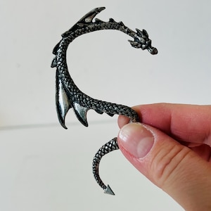 Op de afbeelding: Een zilverkleurige drakenoormanchet. De manchet wikkelt zich om het oor met een drakenkop aan de bovenkant en een staart aan de onderkant. De vleugels en het lichaam van de draak zijn gedetailleerd met schubben en textuurpatronen.