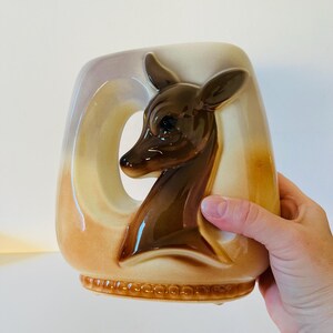 Może przedstawiać: Wazon ceramiczny w stylu vintage z motywem głowy jelenia. Wazon ma brązową głowę jelenia na kremowym i beżowym tle. Uchwyt ma duży, otwarty owalny kształt. Podstawa ma dekoracyjny, koralikowy wz&oacute;r.