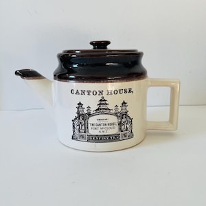 Puede incluir: Una tetera de cerámica marrón y blanca con asa y tapa. La tetera tiene una imagen en blanco y negro de un edificio con el texto "CANTON HOUSE, OBSERVE! THE CANTON HOUSE, FORT MCCLOUD N.W.T. GENUINE TEA" impreso en el lateral.