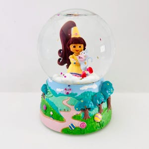 Pode incluir: Um globo de neve decorativo com uma personagem de desenho animado com um vestido amarelo e uma criatura azul. A base é pintada com uma paisagem de caminho, árvores e um castelo. O globo é preenchido com água e glitter.