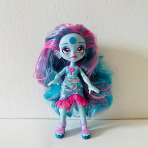 May include: A doll with blue skin, pink and blue hair, and large eyes. The doll wears a blue and pink floral dress with a pink ruffled hem and matching shoes. The doll has a blue dot on its forehead and is posed with arms outstretched.