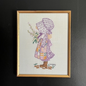 May include: A framed embroidery art piece on white fabric featuring a child in a bonnet and patchwork dress holding flowers. The design includes purple, yellow, and green thread, brown boots, and the embroidered text H. HOBBIE at the bottom.