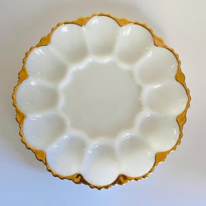 Op de afbeelding: Een ronde witte porseleinen eierplaat met twaalf individuele ovale inkepingen in een cirkel. De geschulpte rand van de plaat is versierd met een metallic gouden bies die de gebogen rand en de buitenste rand van de plaat accentueert.