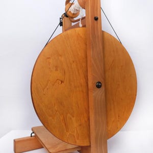 May include: A wooden spinning wheel, featuring a large circular wheel and a smaller spool, crafted from light-toned wood with a natural finish. Black string and white yarn are visible. This wheel is designed for spinning yarn, a traditional craft.