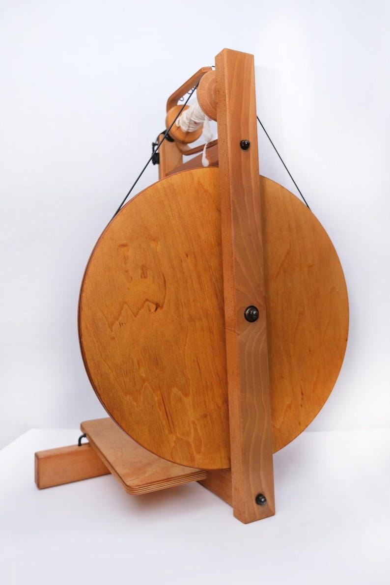 May include: A wooden spinning wheel, featuring a large circular wheel and a smaller spool, crafted from light-toned wood with a natural finish. Black string and white yarn are visible. This wheel is designed for spinning yarn, a traditional craft.