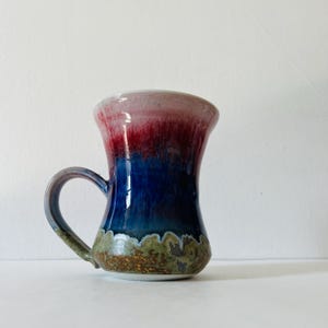 Puede incluir: Una taza de cerámica hecha a mano con un diseño único. La taza presenta un degradado de colores, comenzando con un rojo rosado en el borde, pasando a un azul profundo y terminando con una base texturizada verde y marrón. Tiene un asa curva.