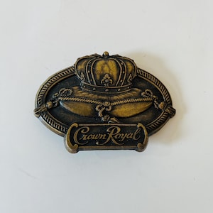 May include: A bronze-colored metal emblem with a crown design and the words "Crown Royal". The emblem is oval-shaped with a rope-like border. The crown is detailed with intricate patterns.