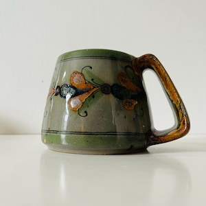Peut inclure: Une tasse en céramique faite à la main avec un design unique. La tasse présente une base vert clair avec une bande décorative de motifs floraux orange, bleus et verts. La poignée a un aspect brun texturé. Convient aux boissons chaudes ou froides.