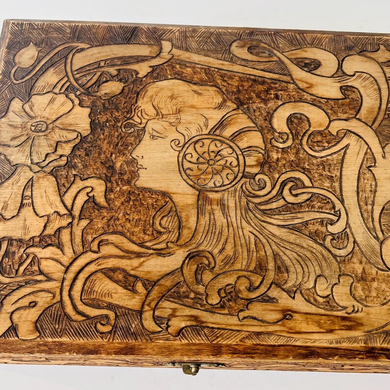 May include: Wooden box with a detailed Art Nouveau style carving. The design features a woman's profile, flowing hair, and floral elements. The box is stained a warm brown color and has a brass clasp.