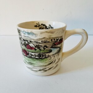 May include: A white ceramic mug with a brown and green country scene design. The scene features a small town with houses, a river, and trees. The mug has a handle and is in good condition.