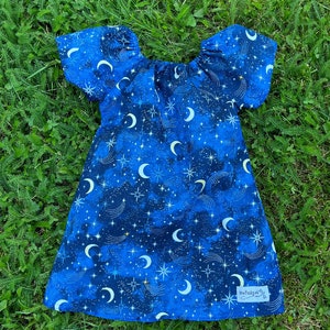 May include: A blue and black dress with a moon and star pattern. The dress has a gathered neckline and short puff sleeves. The dress has a label that says "My Little Sunbeams".