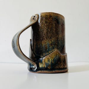 Puede incluir: Una taza cilíndrica de cerámica con un esmalte oscuro y moteado en tonos marrón, negro y azul profundo. La taza tiene un asa ancha y curva unida en la parte superior e inferior, con un pequeño detalle decorativo de cuatro agujeros arriba.