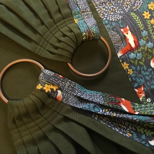 May include: A green fabric baby wrap with a patterned trim featuring foxes and flowers. The wrap has two copper rings.