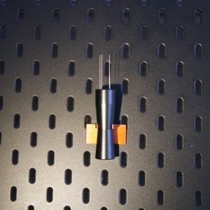 Peut inclure: Un objet en métal noir avec deux tiges métalliques fines s'étendant du haut. L'objet est monté sur une petite base rectangulaire de couleur cuivre.