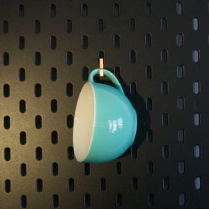 Puede incluir: Una taza de cerámica azul claro con interior blanco cuelga de una pared de tablero de clavijas negro. La taza se mantiene en su lugar mediante un pequeño clip de madera.