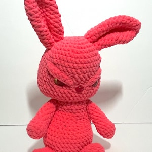Puede incluir: Una muñeca de conejo de ganchillo de color coral hecha a mano con una expresión severa. El conejo tiene orejas largas y erguidas, brazos pequeños y una postura sentada. Los ojos son dorados y la nariz roja. La muñeca está hecha de un hilo suave y texturizado.