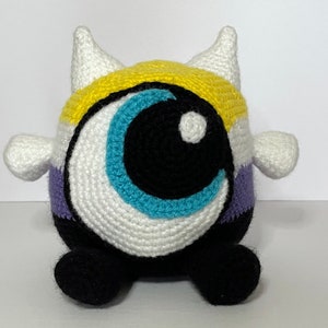Peut inclure: Un jouet en peluche crocheté en forme d'œil avec un schéma de couleurs jaune, blanc, violet et bleu. Le jouet a deux petites cornes sur le dessus et un œil noir.