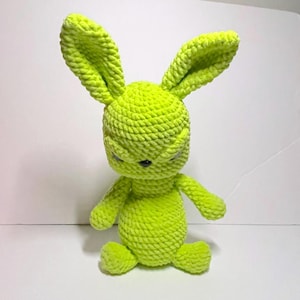 May include: A lime green crocheted bunny doll with long ears, small arms, and a round body. The bunny has a stern expression with closed eyes and a black nose. The plush toy is sitting down.