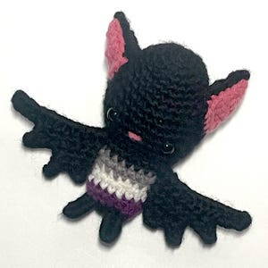 May include: A handmade crochet bat in black with pink and black ears and wings. The bat's belly features the asexual pride flag colors: purple, gray, white, and black. The bat has small black eyes and a pink mouth.