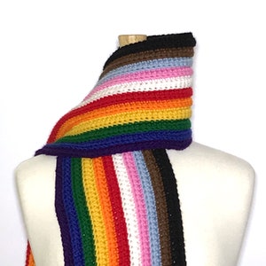 May include: A handmade crochet scarf in a rainbow of colors, including red, orange, yellow, green, blue, and purple. The scarf is displayed on a mannequin and features additional stripes of pink, brown, black, and white.