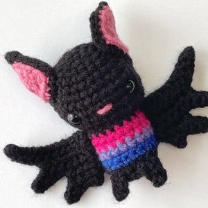 May include: A handmade crochet bat in black yarn with pink and black ears, and a pink mouth. The bat has a colorful collar in pink, purple, and blue. The bat has black safety eyes and outstretched wings.