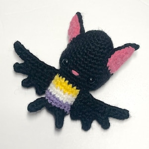 May include: A handmade crochet bat with a black body, pink ears, and a small pink nose. The bat has a non-binary pride flag design on its chest, featuring yellow, white, purple stripes. The bat's wings are spread out.