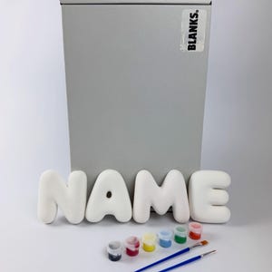 May include: A set of white, rounded, three-dimensional letters spelling the word NAME, positioned in front of a tall, rectangular, silver-coloured box labelled BLANKS. Included are six small paint pots in various colours and two blue paintbrushes.