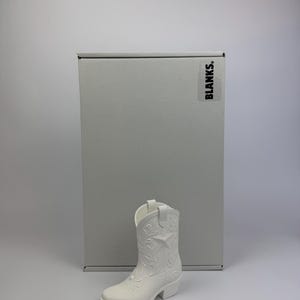 May include: A solid white cowboy boot figurine featuring embossed floral and star patterns on the shaft and toe. The boot is positioned in front of a rectangular grey box with the word BLANKS. printed in black on the upper right corner.