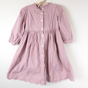 May include: A light mauve dress with long sleeves and a scalloped hem. The dress features a ruffled collar, a pleated bodice, and white buttons down the front. The dress is hanging on a wooden hanger.