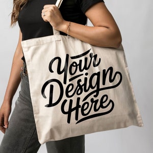 May include: A person is holding a white canvas tote bag with black text that says "Your Design Here".