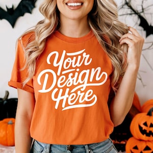 Puede incluir: Una camiseta naranja brillante con el texto "Your Design Here" en blanco. La camiseta está siendo modelada por una persona con cabello largo y rubio. El fondo es una pared blanca con decoraciones de Halloween.