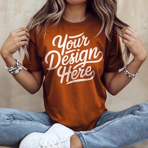 May include: A woman wearing a rust-colored t-shirt with the text "Your Design Here" in white. She is sitting with her legs crossed and wearing blue jeans and white sneakers.