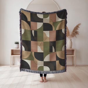 May include: Geometric patterned throw blanket in shades of olive green, black, tan, and blush pink. The design features a repeating pattern of semi-circles, lines, and squares. The blanket has a fringed edge.