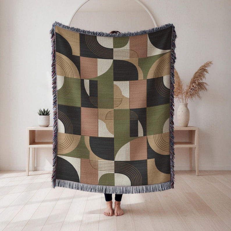May include: Geometric patterned throw blanket in shades of olive green, black, tan, and blush pink. The design features a repeating pattern of semi-circles, lines, and squares. The blanket has a fringed edge.