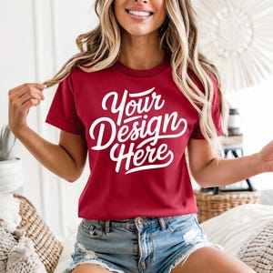 May include: A maroon t-shirt with the text "Your Design Here" in white script. The short-sleeved shirt is paired with denim shorts. The model is smiling and holding their hair.