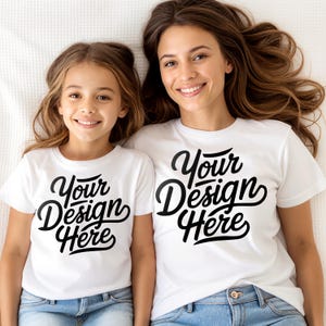 Könnte beinhalten: Zwei weiße T-Shirts mit dem Text "Your Design Here" in schwarzer Schrift. Die Shirts werden von einer Frau und einem Kind getragen, beide lächeln. Die Shirts werden mit blauen Jeans kombiniert.