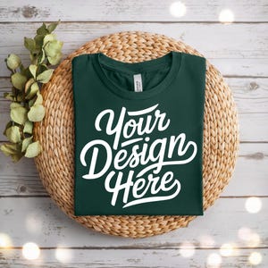 May include: A dark green t-shirt with the text "Your Design Here" in white script. The shirt is neatly folded and placed on a round woven mat, with sprigs of greenery to the left. The background is a light wood surface.