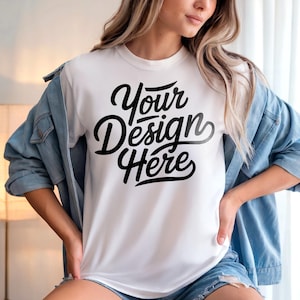 以下が含まれることがあります： 黒い筆記体フォントで「Your Design Here」というテキストが入った白いTシャツ。明るいブルーのデニムジャケットと合わせて着用。モデルはデニムのショートパンツも着用しています。
