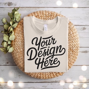 May include: Cream-colored t-shirt with the text "Your Design Here" in black script. The shirt is folded and placed on a round woven mat, with greenery and decorative lights in the background. The shirt is a blank canvas for custom designs.