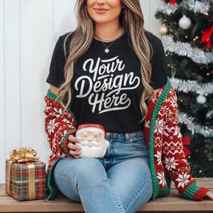 May include: Black t-shirt with the text "Your Design Here" in white script. The person is wearing a red and white patterned sweater and holding a Santa Claus mug. A wrapped gift box with a gold bow is on the left.