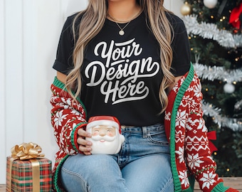 May include: Black t-shirt with the text "Your Design Here" in white script. The person is wearing a red and white patterned sweater and holding a Santa Claus mug. A wrapped gift box with a gold bow is on the left.