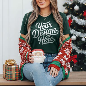 May include: A dark green t-shirt with the text "Your Design Here" in white script. The person is wearing a red and white snowflake patterned cardigan and holding a Santa Claus mug. A wrapped gift box with a gold bow is on the left.