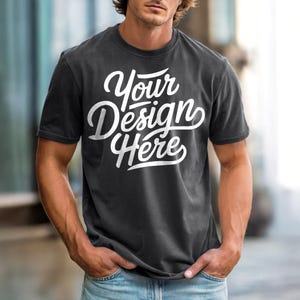 May include: Dark gray t-shirt with the text "Your Design Here" in white script. The shirt has short sleeves and a crew neck. The model is wearing blue jeans.