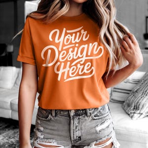 Puede incluir: Una camiseta naranja brillante con el texto "Your Design Here" en letras blancas. La camiseta está hecha de una tela suave y cómoda y tiene un corte holgado.