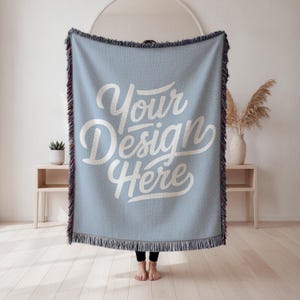 May include: A light blue woven throw blanket with the text "Your Design Here" in white script. The blanket has a fringed edge with a mix of colors. The blanket is displayed indoors.
