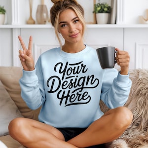 Może przedstawiać: Jasnoniebieska bluza z napisem "Your Design Here" w czarnym skrypcie. Osoba trzyma bluzę i czarny kubek. Osoba siedzi na kanapie.