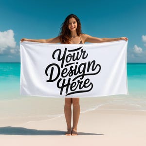 May include: White rectangular beach towel with the text "Your Design Here" in black script. The towel is held up against a backdrop of a beach with turquoise water and a blue sky.