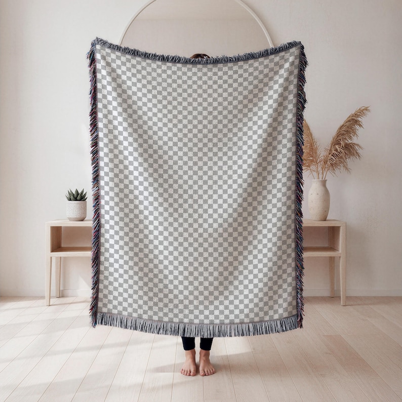 May include: A large, checkered throw blanket in shades of gray and white, with a fringed edge. The blanket is held up, showcasing its pattern and texture. The throw is a home decor item.